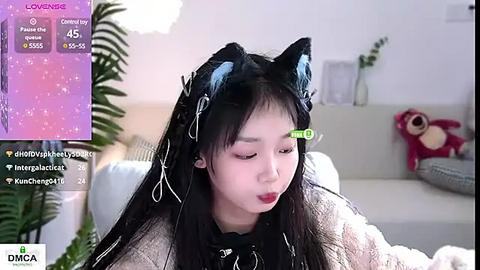 Media: A video of an East Asian woman with long black hair, wearing a black cat headband, red lipstick, and a beige sweater, seated on a white couch in a light room.