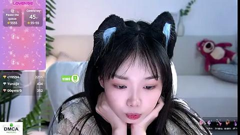 Media: Video of a young East Asian woman with long black hair and cat ears, resting her chin on her hands, in a modern, minimalist room with a pink teddy bear.