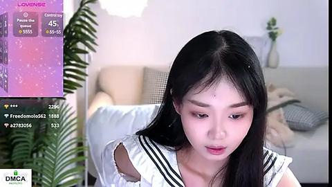 Media: Video of an East Asian woman with long black hair, wearing a white sailor-style school uniform, sitting indoors. Background includes a beige couch and potted plants.