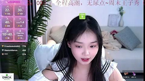 Media: Video of an East Asian woman with long black hair, wearing a white sailor-style top, sitting in a cozy living room with a gray couch, flowers, and a green plant.