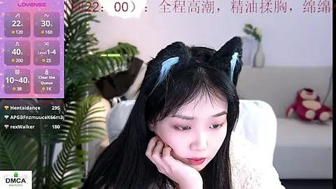 Media: A video of a young Asian woman with long black hair, wearing cat ears, seated indoors with a plush toy and a white couch in the background.