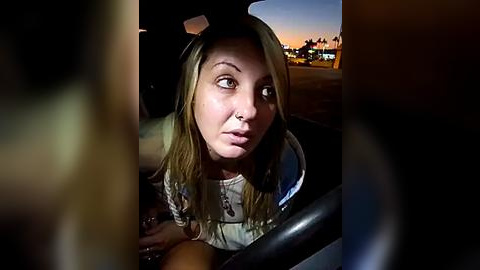 Media: Video of a blonde woman in a car, looking contemplative at twilight. She wears a beige top, with a blurred urban cityscape visible through the window.