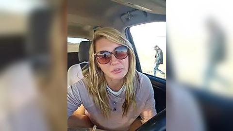 Media: A video of a blonde woman with long hair, wearing sunglasses, a light gray shirt, and a necklace, sitting in a car, blurred background.