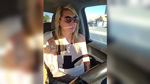 Media: A video of a blonde woman with tattoos, wearing sunglasses, seated in a car with a sunlit view of a suburban street and a gas station.