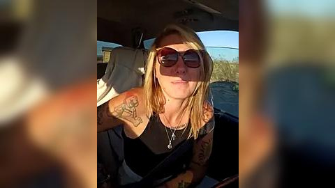 Media: Video of a blonde woman with tattoos, wearing dark sunglasses, driving a car, with a blurred background featuring desert scenery and a clear blue sky.