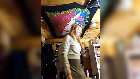 Media: Video of a blonde woman in a beige sweater and green skirt, walking in a cluttered, rustic room with a vibrant, abstract mural on the ceiling and wooden walls.