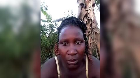 Media: Video of a dark-skinned woman with braided hair, wearing a beige bra, standing outdoors in front of a palm tree. The background features lush green foliage and a blurred, out-of-focus setting.