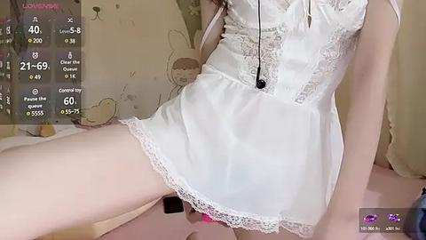 Media: A video of a fair-skinned person in a white, lace-trimmed babydoll dress, sitting on a bed. Background shows a wall with cartoonish bunny patterns.