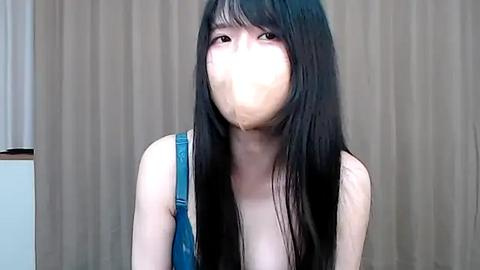 Media: A video of an Asian woman with long, straight black hair, wearing a blue tank top, sitting against beige curtains. She has a white mask over her mouth, giving a mysterious and slightly eerie vibe.