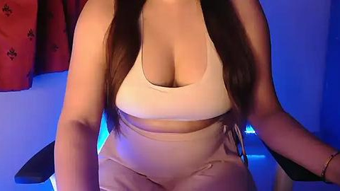 Media: Video of a curvy woman with long, straight brown hair, wearing a tight, white crop top that reveals ample cleavage and high-waisted beige pants. She sits on a chair with a red and black patterned cushion in a dimly lit room with blue lighting.