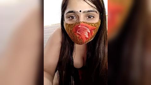 Media: Video of a woman with long, dark hair, wearing a red and brown floral face mask, with a white dot on her forehead, against a blurred background.