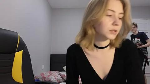 Media: Video of a blonde woman in a black V-neck top, standing in a minimalist bedroom with a black gaming chair, a bed with floral sheets, and another person in the background.