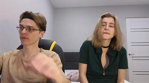 Media: Video of a young man with glasses and short brown hair in a beige T-shirt, and a young woman with blonde hair and a black choker, both sitting in a modern, minimalist room with grey walls.