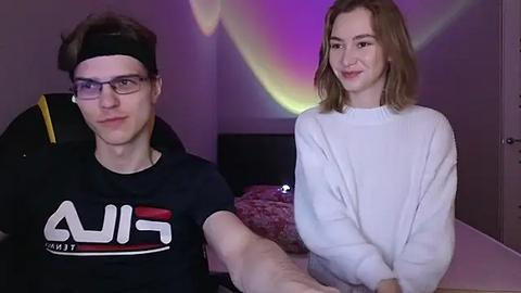 Media: Video of a young Caucasian man with glasses, black T-shirt, and headphones, and a young Caucasian woman with shoulder-length blonde hair, white sweater, in a dimly lit room with purple and yellow light.