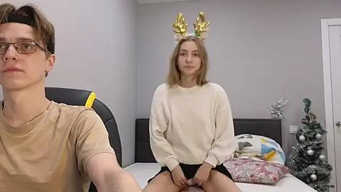 Media: A young woman with blonde hair and antlers, wearing a beige sweater, sits on a bed. A man with glasses and short hair sits beside her.