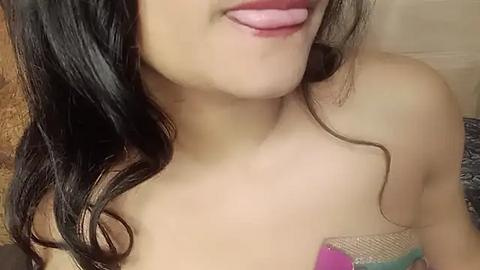 Media: A close-up video of a young woman with light brown skin, dark wavy hair, and pink lipstick. She is topless, showing her bare shoulders. The background features a textured, beige-colored wall.