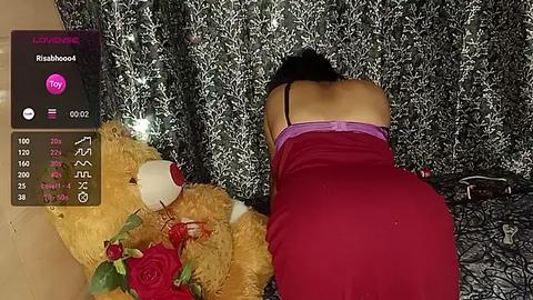 Media: A video captures a woman in a red dress kneeling on a patterned floor, leaning on a large stuffed teddy bear adorned with a red rose. The background features a floral-patterned curtain.