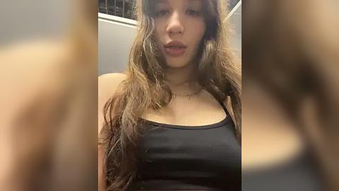 Media: Video of a young woman with long, wavy brown hair, wearing a black tank top, standing indoors with blurred background.