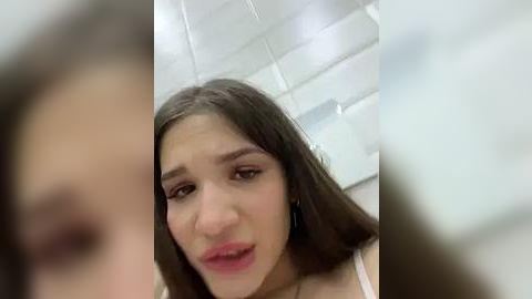 Media: Video of a young woman with straight, shoulder-length brown hair, fair skin, and light makeup, looking concerned or confused. She's in a bathroom with white tiled walls, blurry in the background.