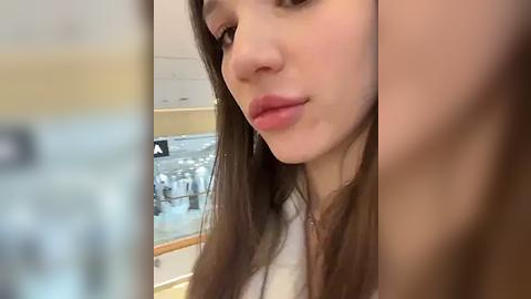 Media: Video of a close-up of a young woman with fair skin and long brown hair, wearing a white shirt, in a modern kitchen with stainless steel appliances and glass cabinets.