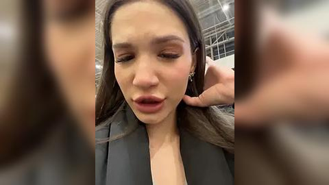 Media: Video of a young woman with long brown hair, wearing a grey blazer, adjusting her earring with a smile, in an indoor setting with blurred background.