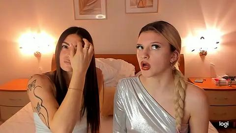 Media: Video of two young women in a bedroom. One with long dark hair, the other with blonde braided hair, both wearing metallic dresses, looking surprised. Bed with white sheets, bedside lamps, and framed art on beige walls.