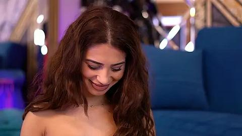 Media: Video of a young woman with long, wavy brown hair, light skin, and a slender figure, smiling softly, wearing a strapless top. She's seated on a blue couch in a dimly lit room with bokeh lights in the background.