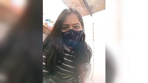 Media: Video of a young woman with long, dark hair and a black and white striped shirt, wearing a blue face mask, taken from a slightly blurred angle, indoors, with a tiled ceiling.