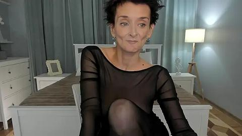 Media: Video of a middle-aged woman with short dark hair, wearing a sheer black top, sitting in a modern, minimalist bedroom with white furniture, gray curtains, and a lamp.