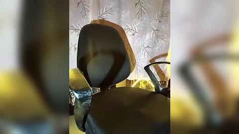 Media: Video of a black motorcycle helmet with a brown visor, placed on a light brown leather chair with a yellow cushion, against a backdrop of white, floral-patterned curtains. The image is slightly blurred.