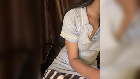 Media: Video of a young woman with medium brown skin, wearing a light blue polo shirt and striped shorts, sitting on a dark brown leather chair, with dark curtains in the background.