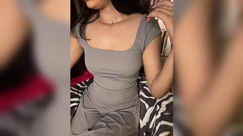 Media: Video of a woman with medium skin tone, black hair, wearing a tight gray t-shirt and high-waisted pants, sitting on a zebra-patterned bed, holding a phone, blurred background.