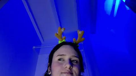 Media: A video of a young woman with fair skin, dark hair, and a small nose, wearing a green headband with yellow reindeer antlers and a white flower, set against a blue-lit ceiling.