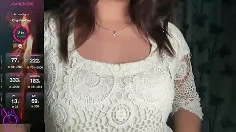 Media: Video of a woman with medium brown skin, wearing a white lace top, standing indoors. She has long dark hair and a thin gold necklace. The background is blurred.