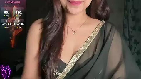 Media: A video of a woman with long dark hair, wearing a black saree with gold embroidery, smiling slightly. Background shows a dimly lit room with a TV displaying \"Live\" and \"Loknase.\