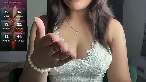 Media: Video of a woman with medium skin tone, wearing a white lace bra, making a shaka sign with her right hand. She has long, dark hair, and a pearl bracelet on her wrist. Background includes a TV screen displaying a weather forecast.