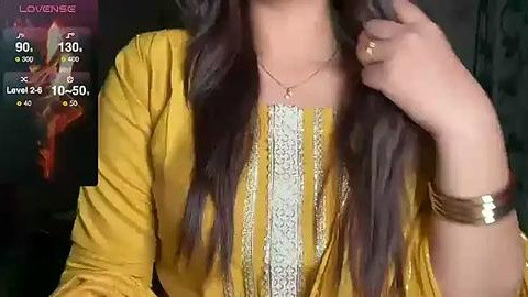 Media: Video of a woman with long, straight brown hair, wearing a mustard yellow kurta with intricate white embroidery, a gold bracelet, and a delicate necklace. The background features a blurred, dark setting with a \"Lovenee\" logo and digital stats in the corner.