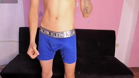 Media: Video of a fit, shirtless man with fair skin wearing tight blue Calvin Klein boxer briefs and a sleeve tattoo, standing against a pink wall with snowflake stickers, on a black couch.