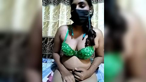 Media: Video of a curvy, South Asian woman with medium brown skin, wearing a green lace bra and black mask, sitting on a bed with a patterned sheet, in a dimly lit room with floral curtains.