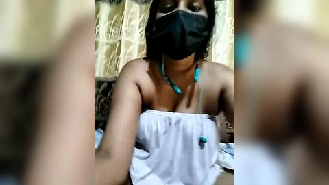 Media: Video of a dark-skinned woman with short hair, wearing a black face mask, turquoise necklace, and a white strapless dress, seated against a beige textured wall. The image is slightly blurry.