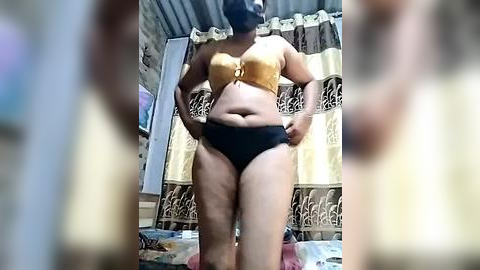 Media: Video of a plus-sized woman in a mustard yellow bra and black panties, standing in a dimly lit, cluttered room with a floral curtain and a bed.