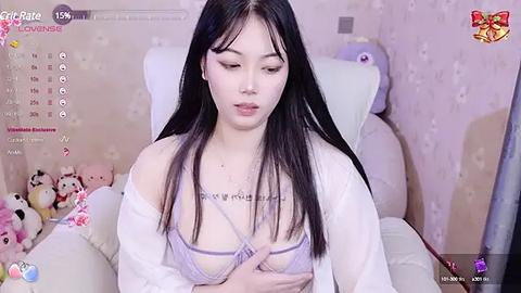 Media: A video of an East Asian woman with long black hair, wearing a white robe and a revealing lavender bra, sitting on a plush chair in a pastel-themed room with plush toys.