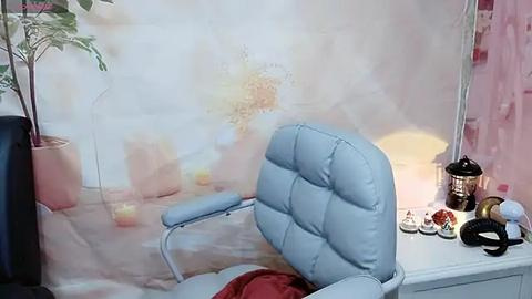 Media: A video of a light blue, cushioned office chair positioned in front of a white desk with a pink floral curtain, a candle, and a potted plant.
