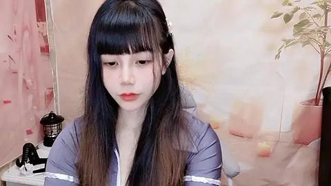 Media: Video of a young Asian woman with long, straight black hair and bangs, wearing a lavender blouse. She has pale skin and wears red lipstick. Background shows a potted plant and abstract artwork.