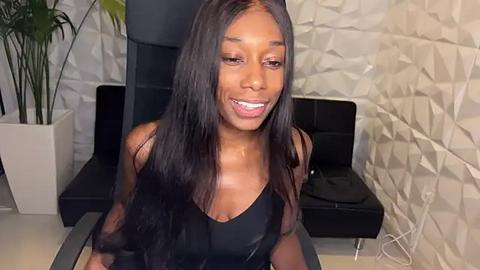 Media: Video of a smiling Black woman with long, straight black hair, wearing a black tank top, sitting in a modern office chair with a textured white wall and black leather cushion behind her.