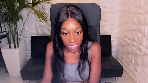 Media: Video of a young Black woman with long straight black hair, wearing a grey tank top, sitting on a black leather chair against a white textured wall.