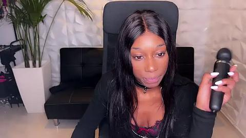 Media: Video of a black woman with long straight hair, wearing a black top and a black choker, holding a black vibrator, sitting in a black leather chair in a modern, minimalist room.