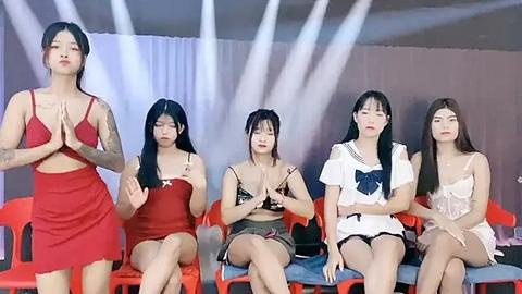 Media: Video of five Asian women, seated, in red and white lingerie, hands clasped, on plastic chairs against a white backdrop, with bright lights.