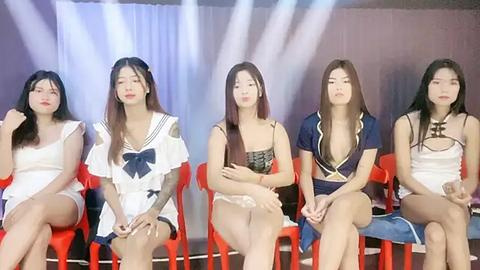 Media: Video of five East Asian women, seated on red plastic chairs, wearing various summer outfits, against a backdrop with white curtains and bright stage lights.