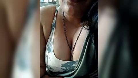 Media: Video of a woman with light skin and dark hair, wearing a low-cut, green and white tie-dye tank top, revealing cleavage, and a silver chain necklace.